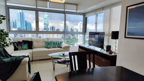 A modern living room with a sofa, coffee table, and dining table. Large windows offer a view of a city skyline. - Beautiful Apt, Best Location 2BR, Best Location (Panamá)