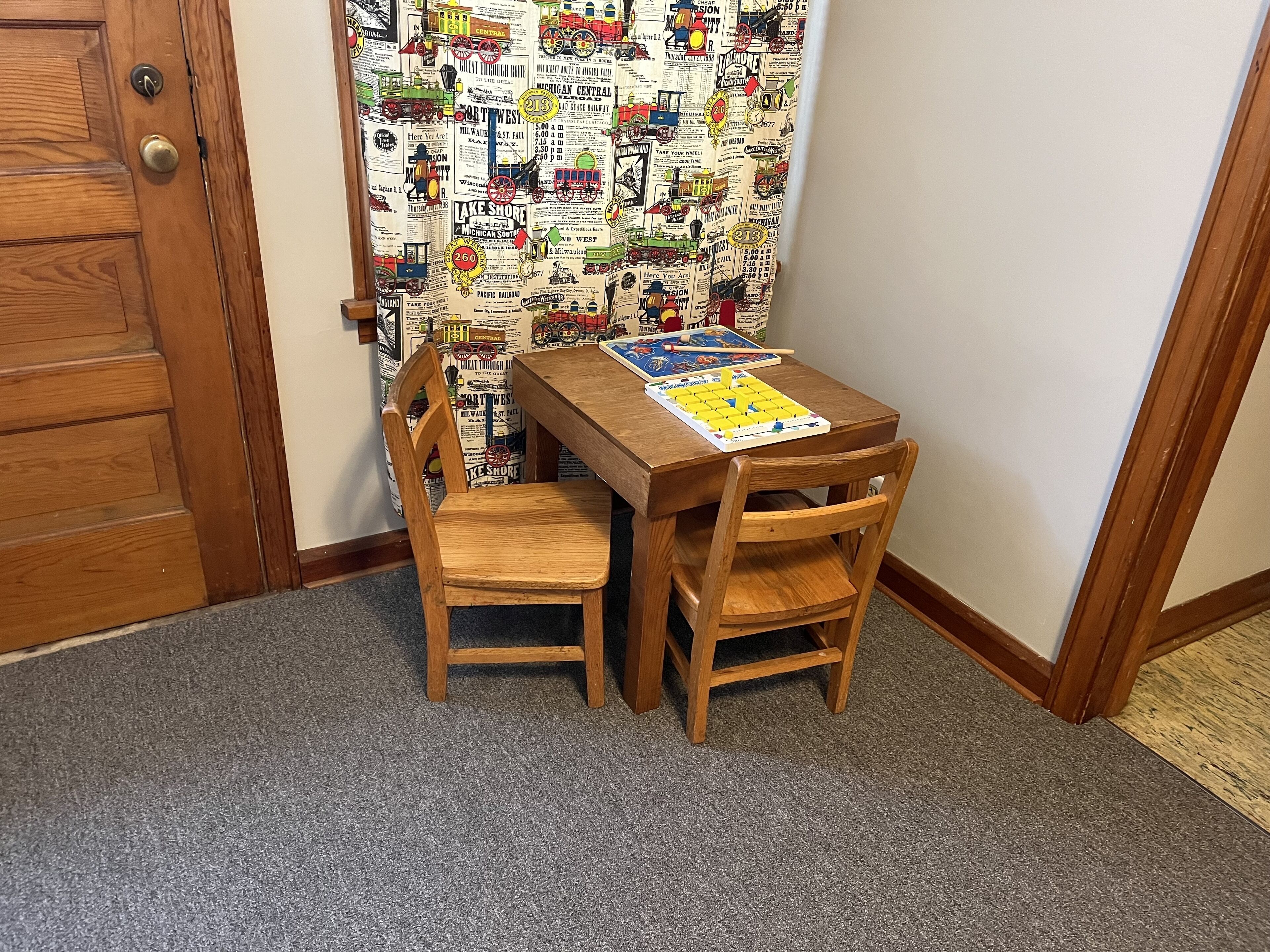 Ein Eckzimmer mit Holztür, Wand mit Muster, ein kleiner Holztisch mit einem Brettspiel und zwei Holzsesseln.