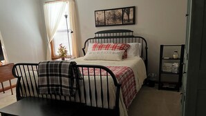 Une chambre à coucher avec un lit, une chaise, une table de chevet, une lampe, un petit sapin de Noël et un tableau encadré accroché au mur.