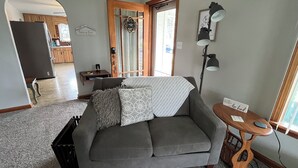 Un salon avec un canapé gris, une petite table d’appoint en bois, une lampe et une porte donnant sur la cuisine.