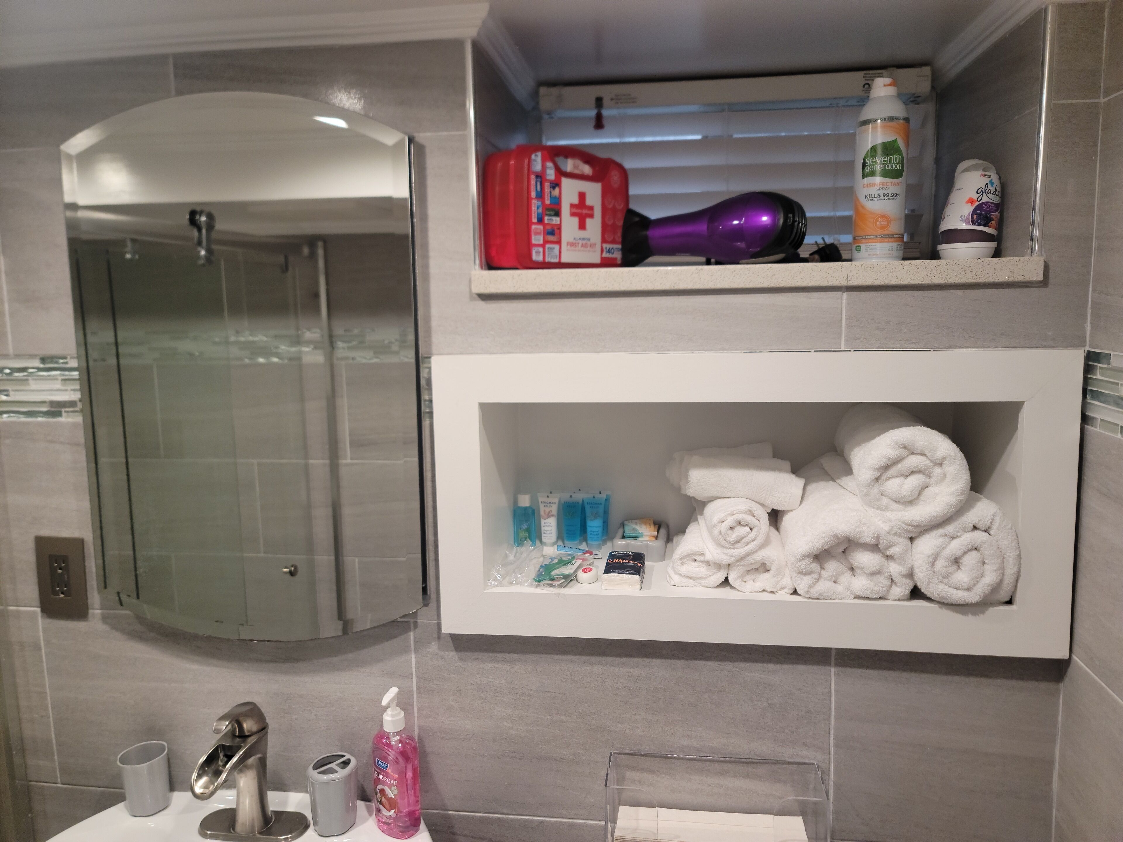 Un bany amb dutxa de vidre, un lavabo amb mirall i prestatges amb productes de tocador i tovalloles.