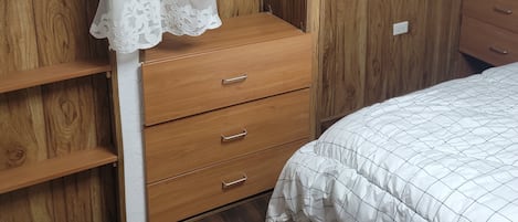 A bedroom with a wooden wardrobe, a bed with a white quilt, and a hanging wardrobe with a white curtain.