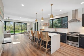 Uma cozinha moderna com uma ilha grande, banquetas, eletrodomésticos de aço inoxidável e janelas amplas com vista para o exterior.