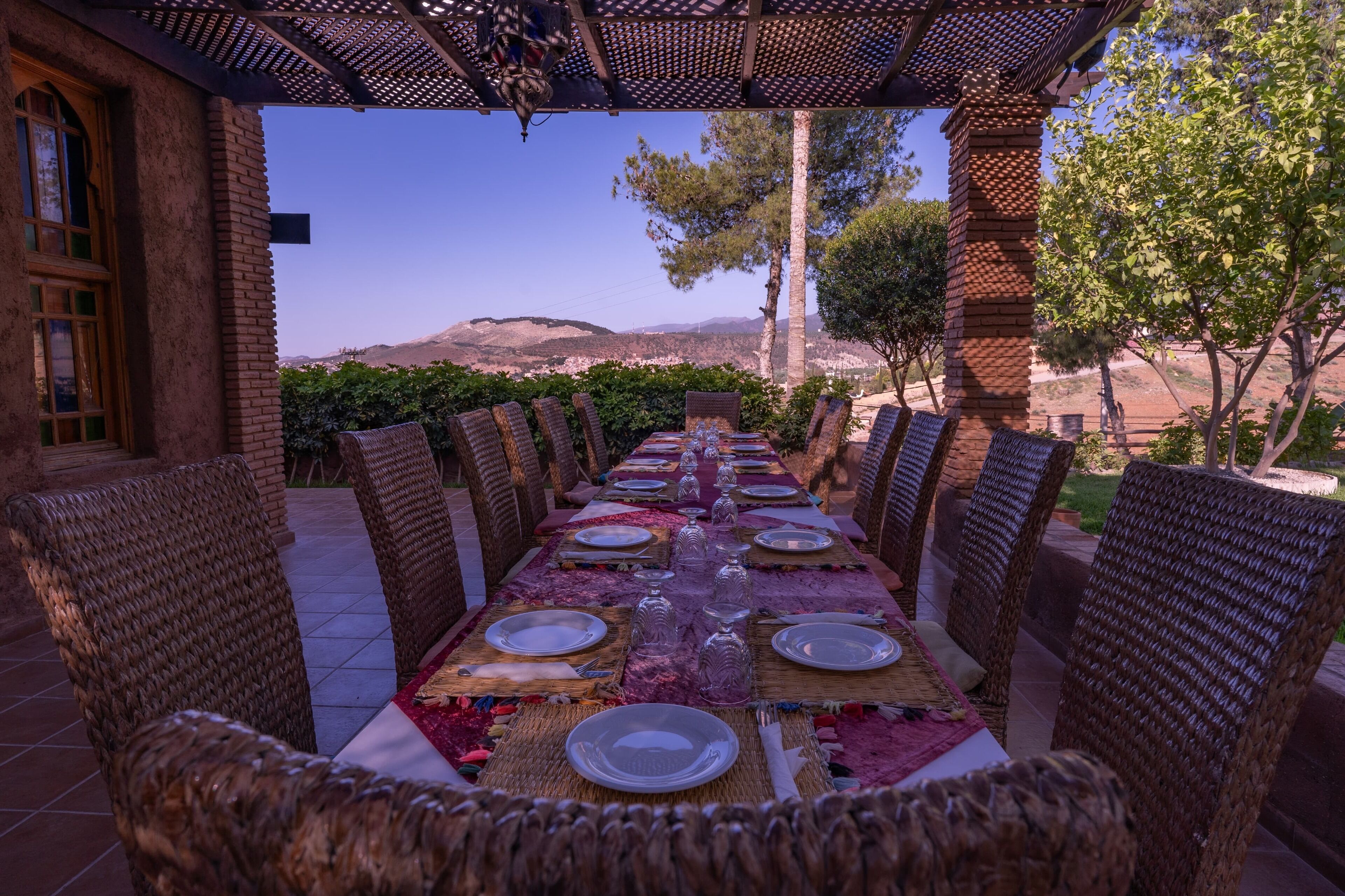 An outdoor dining area with a long table set for a meal, surrounded by wicker chairs, and overlooking a scenic landscape with hills and trees.