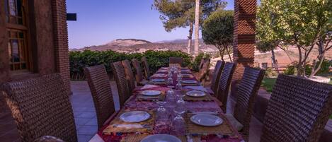 An outdoor dining area with a long table set for a meal, surrounded by wicker chairs, and overlooking a scenic landscape with hills and trees.