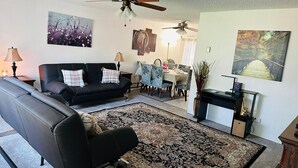 Un salon avec un canapé noir, un tapis à motifs, un ventilateur de plafond et des œuvres d’art accrochées aux murs.