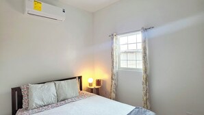 A bedroom with a bed, a ceiling fan, an air conditioning unit, a bedside table with a lamp, a window with curtains, and a patterned rug.