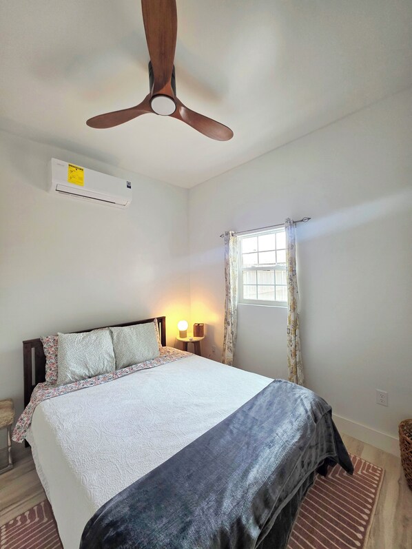 A bedroom with a bed, a ceiling fan, an air conditioning unit, a bedside table with a lamp, a window with curtains, and a patterned rug.