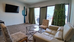 Ein Wohnzimmer mit einer Couch, Sesseln, einem Glastisch, einer Kakteenpflanze und einem an der Wand befestigten Fernseher.