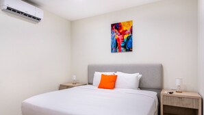 Una habitación de hotel moderna con una cama grande, dos mesitas de noche, un equipo de aire acondicionado y un cuadro abstracto y colorido en la pared.