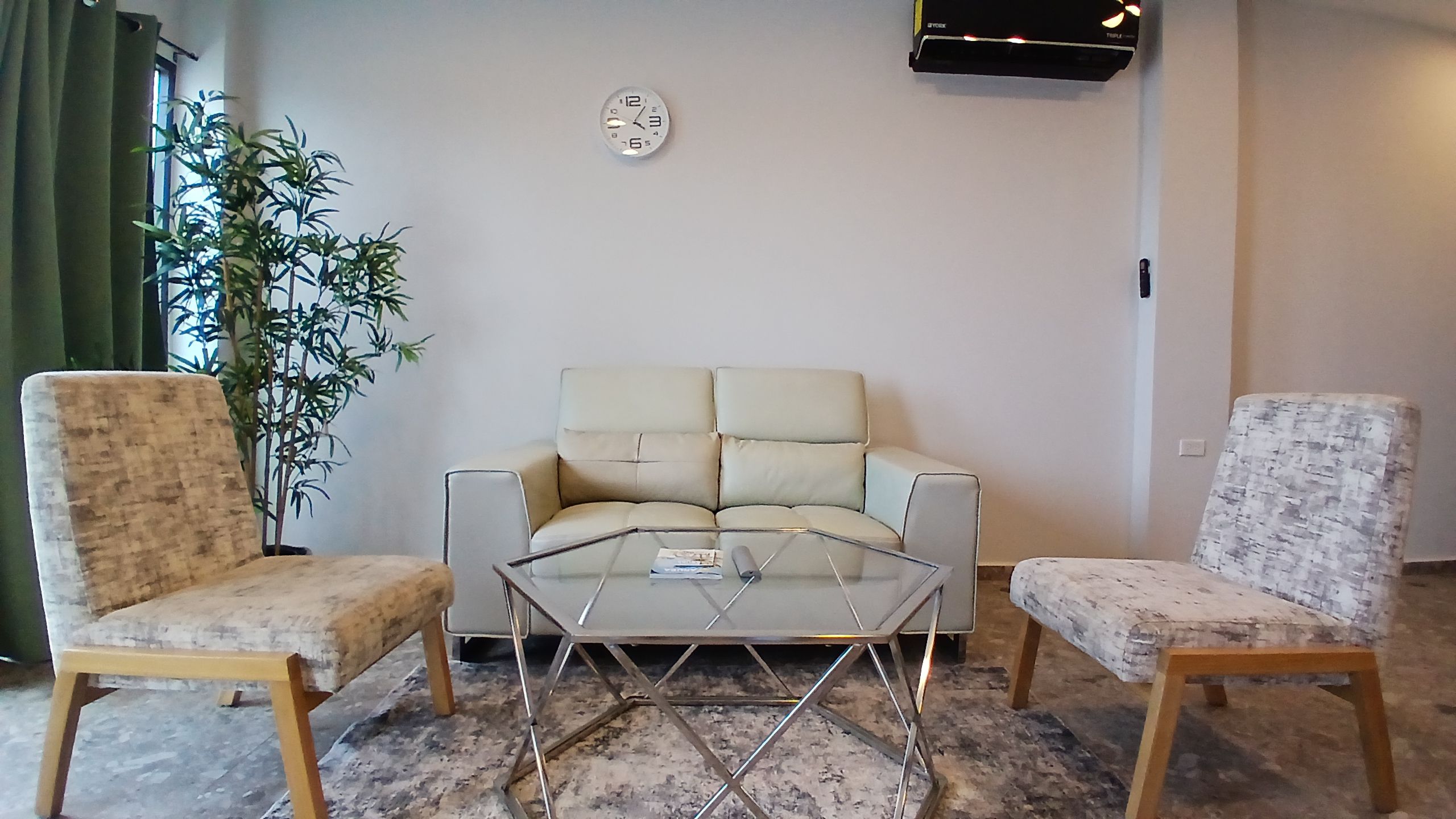 Una sala de estar con un sofá, dos sillones, una mesa de centro de vidrio, un reloj de pared y una planta en maceta.