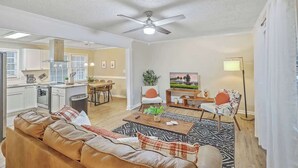 A modern living room with a brown leather sofa, a coffee table, and a TV. There is a kitchen area with white cabinets and a dining table. - Family Fun near CHAS Summerville and Nexton (Summerville)