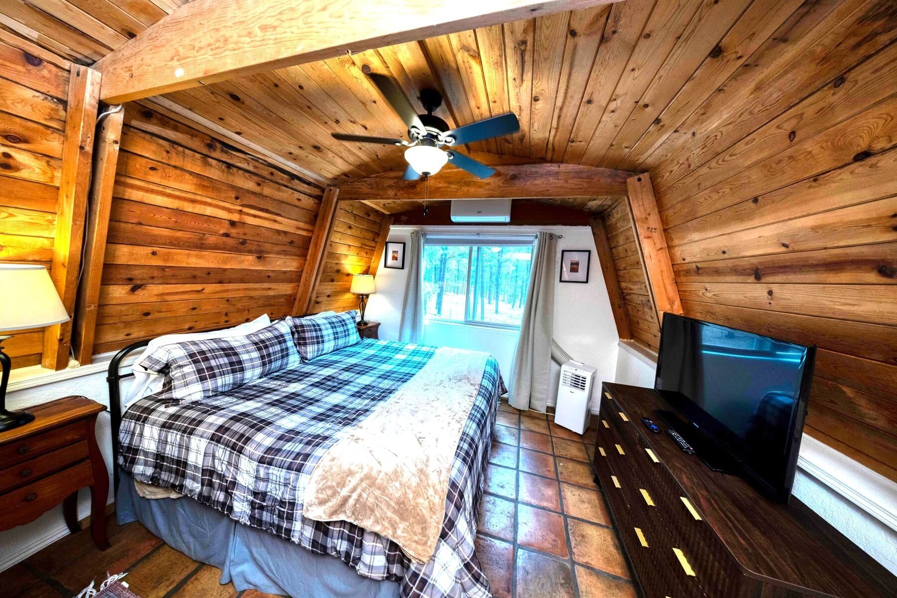 Een gezellige slaapkamer in een chalet, met een bed, plafondventilator, televisie en een raam met gordijnen.