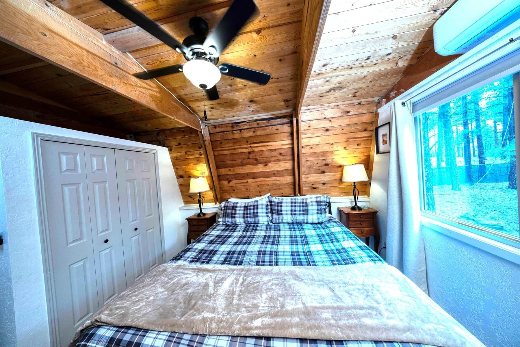 Un dormitorio con cama, ventilador de techo, ventana con vistas a árboles y una puerta.