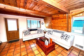 A cozy cabin interior with a wooden ceiling, a sofa set, a coffee table, and a window with a view of a snowy landscape. - Grand Gateway Cabin | 1-Acre Pine Retreat, Rt. 66 (Williams)
