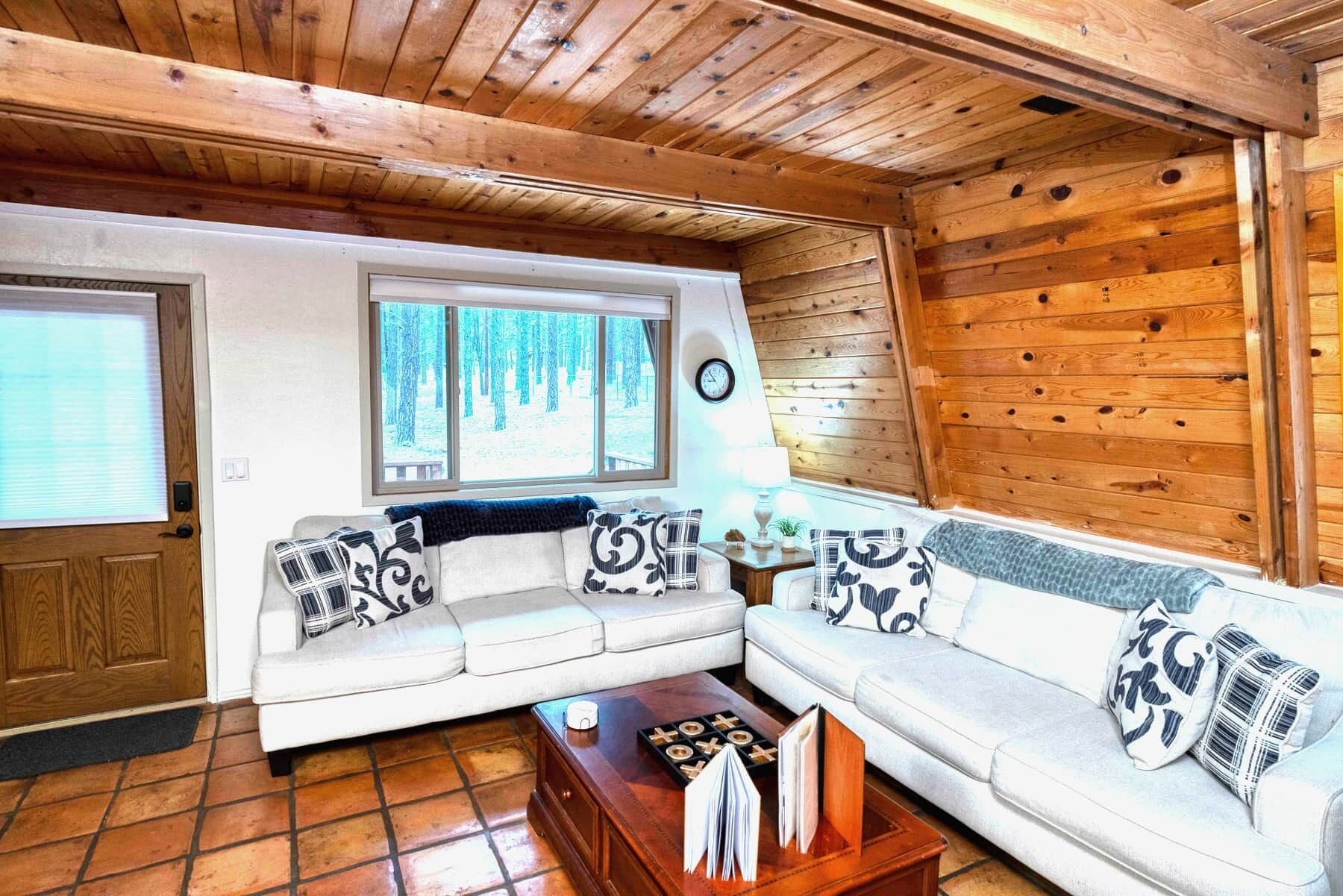 Interior de una cabaña con un sofá blanco, techo de madera, ventana con vistas a árboles y un reloj en la pared.