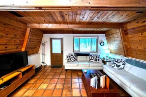 Ein gemütliches Wohnzimmer mit einer Couch, einem Couchtisch, einem Fernseher und einem Fenster mit Blick auf Bäume.