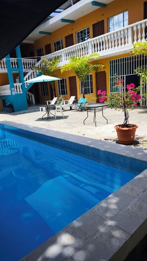 A swimming pool with lounge chairs and a table, surrounded by a building with balconies and potted plants. - Hotel y Restaurante Casa Jardines (San Benito)