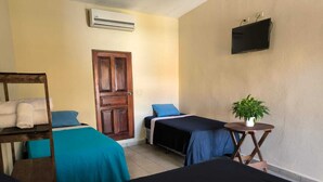A room with two beds, a wooden shelf with towels, a small table with a plant, a TV on the wall, and an air conditioning unit. - Hotel y Restaurante Casa Jardines (San Benito)