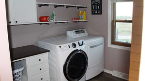 A laundry room with a washing machine, a dryer, a shelf with laundry supplies, and a window with a view. - Brigham View (Barneveld)