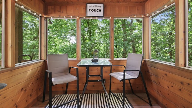 A wooden porch with a table and two chairs, a sign reading "Welcome to the Porch," and large windows offering a view of trees.
