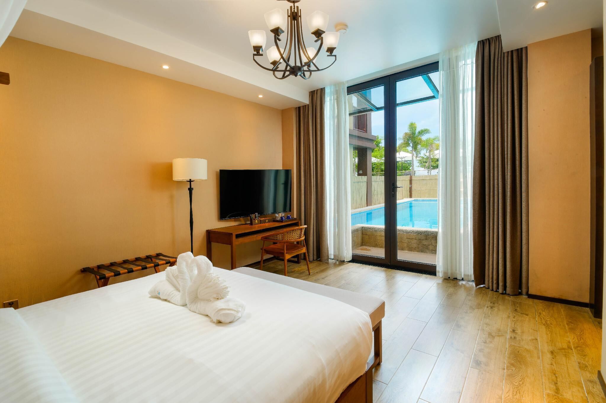 A hotel room with a bed, a desk with a TV, a chair, a robe, and a view of a pool through a glass door.