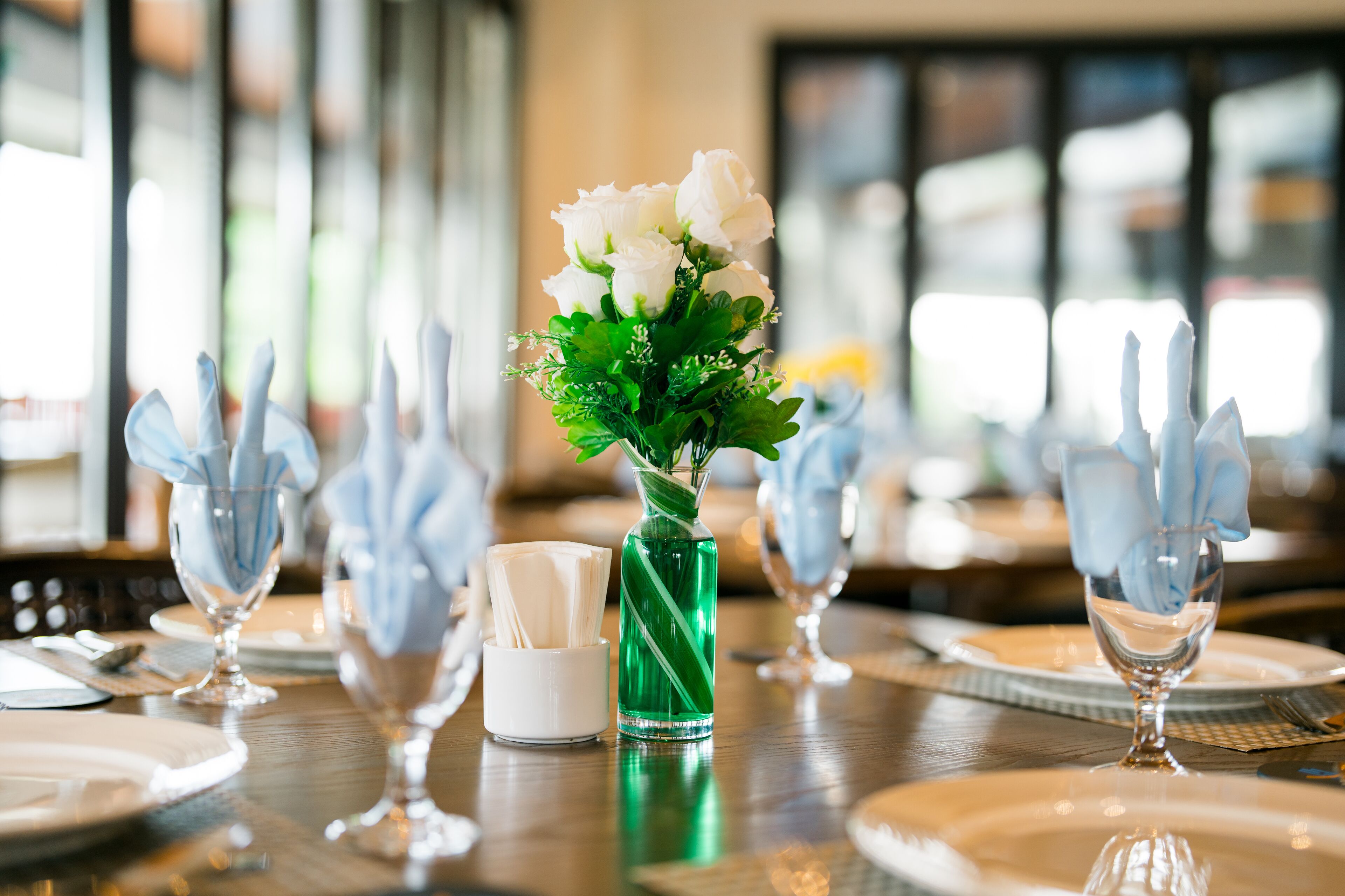 A dining table set with white napkins, a green vase with white flowers, and a white cup.