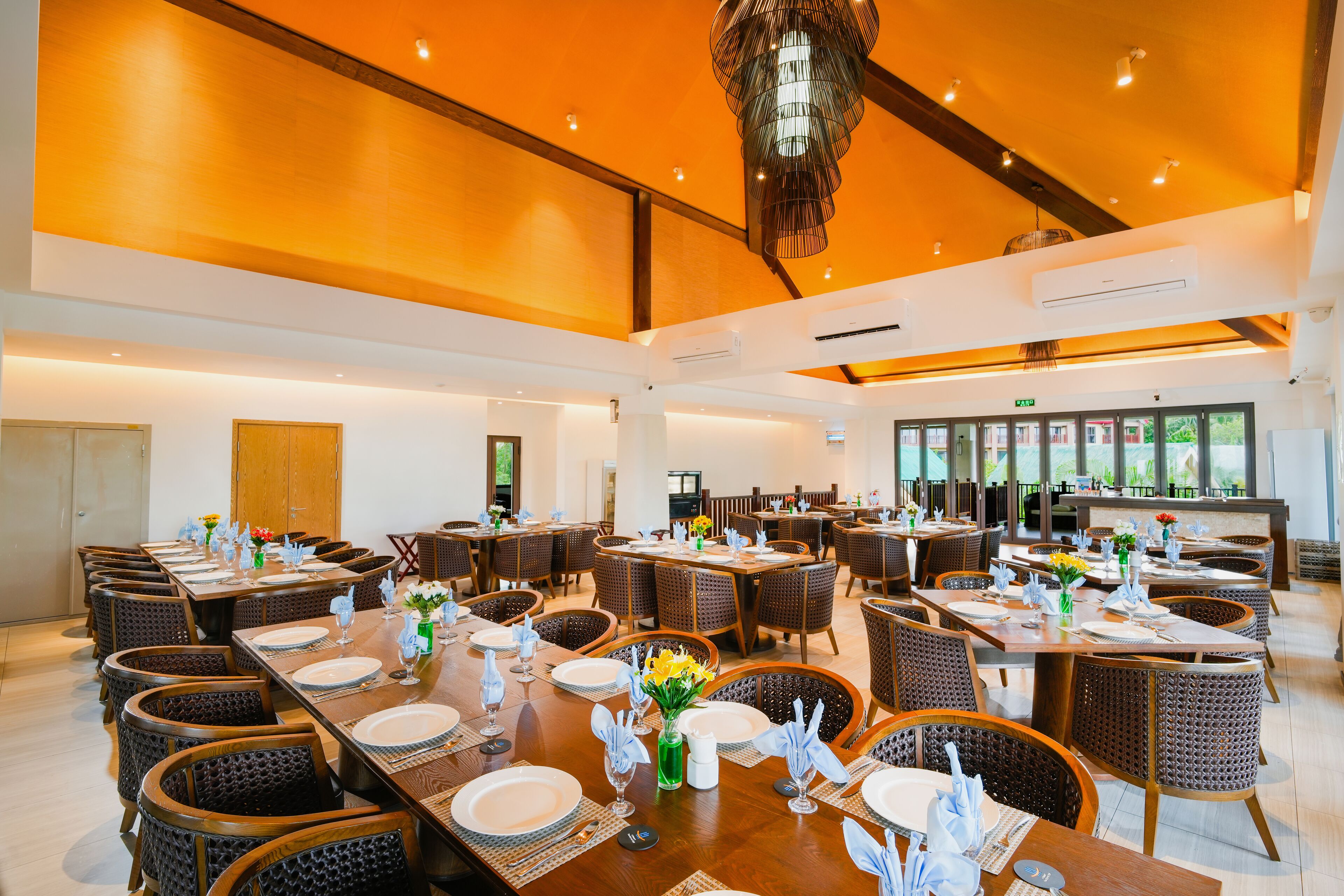 Un vaste espace dédié à la salle à manger, avec de nombreuses tables dressées, des chaises en osier et un lustre moderne.