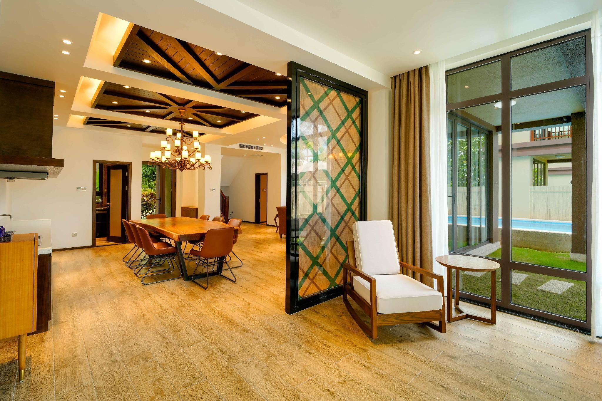 A modern interior with a wooden dining table, chairs, a chandelier, and large windows offering a view of the outdoors.