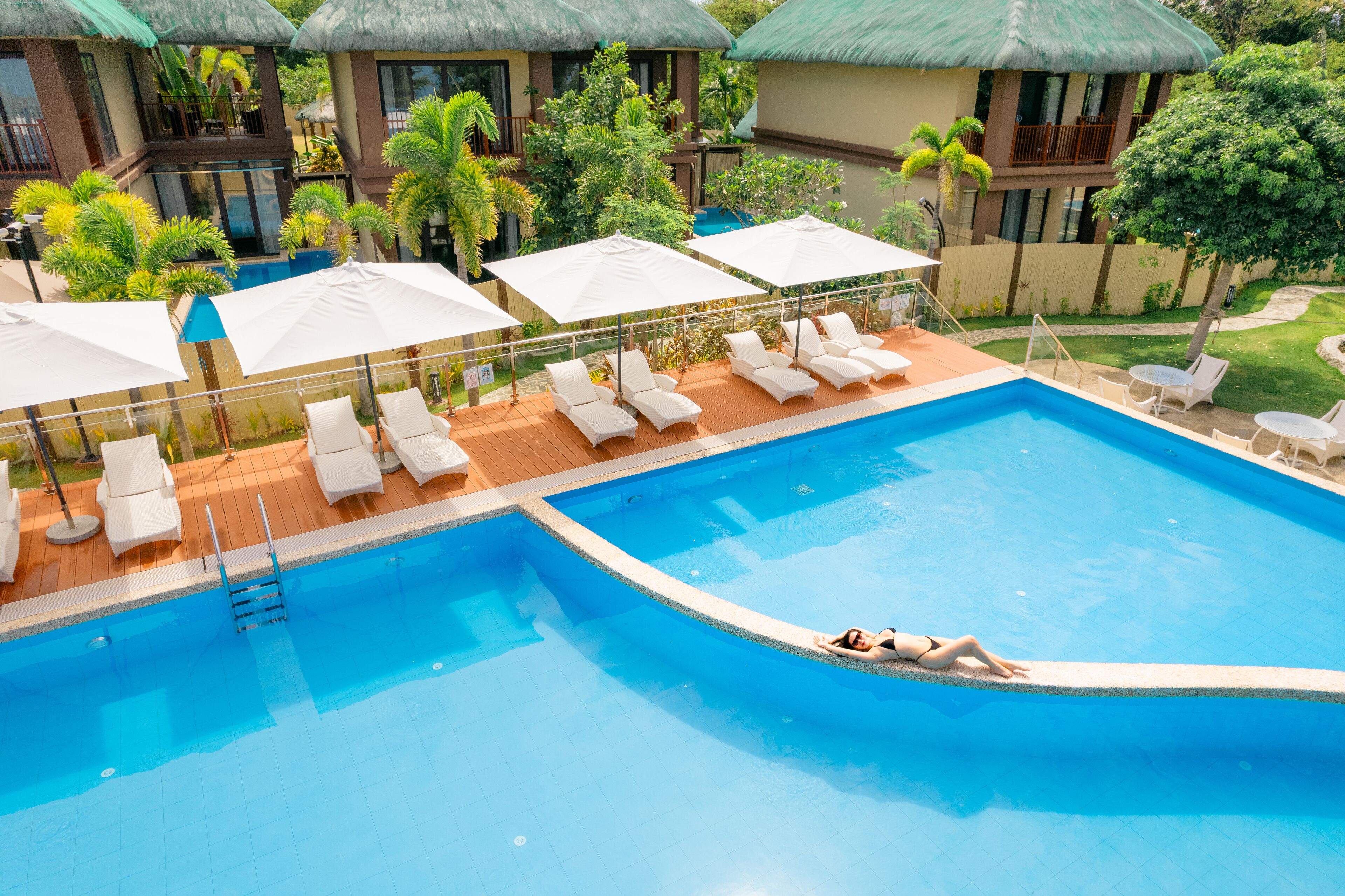 A swimming pool with lounge chairs and umbrellas, surrounded by greenery and buildings.