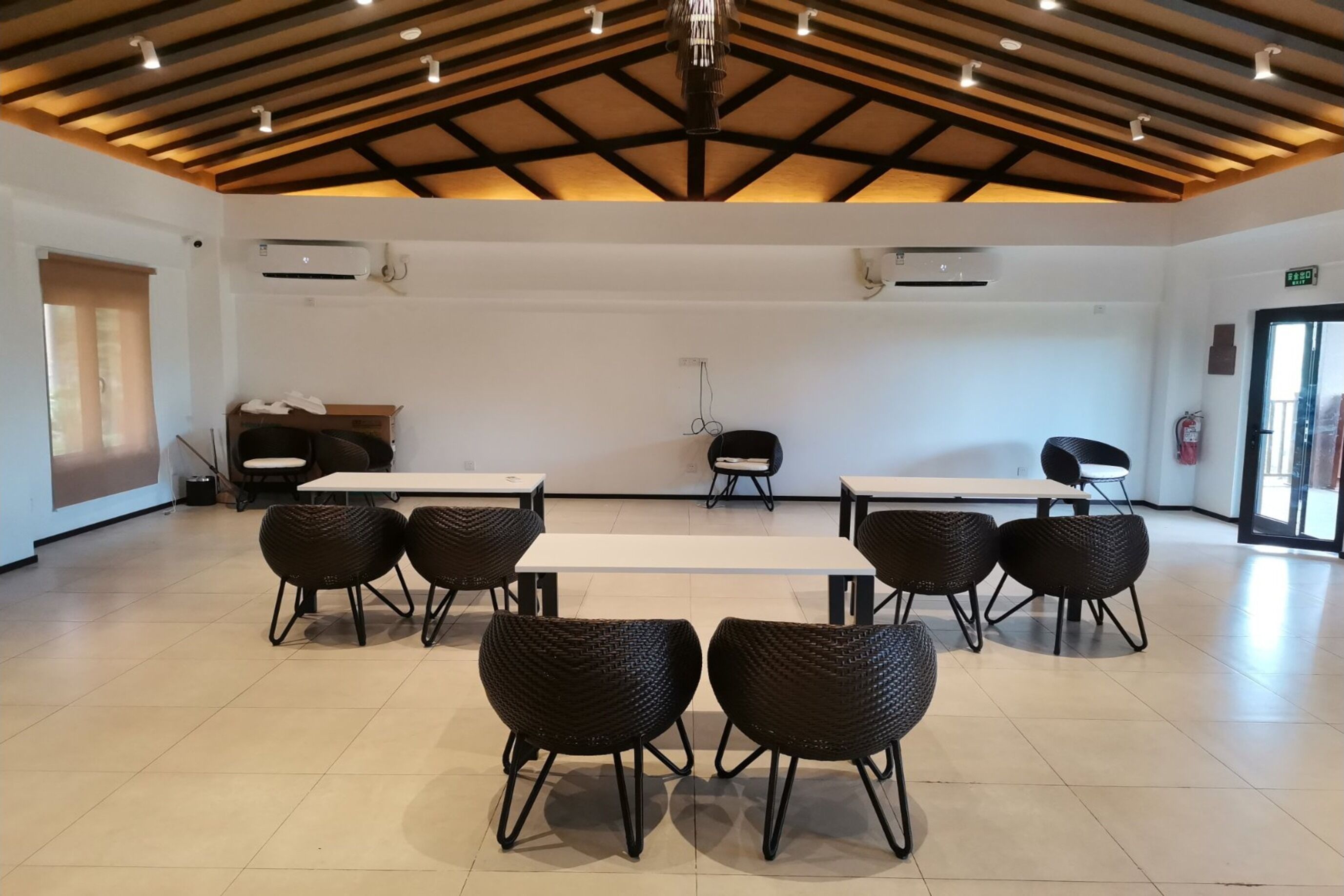 A spacious room with a high ceiling, white walls, and a wooden beam structure. There are several tables and chairs arranged for dining or meetings.
