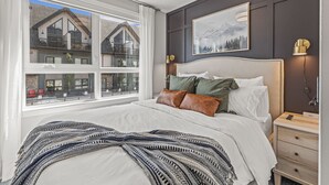 Un dormitorio con una cama grande, una mesita de noche, una lámpara de pared y una ventana con vistas a una casa.