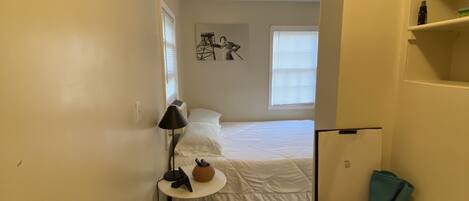 Habitación pequeña con cama, lavadora, una mesita con lámpara y estante en la pared.