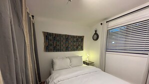 A bedroom with a bed, a nightstand with a lamp, a wall clock, and a window with blinds.
