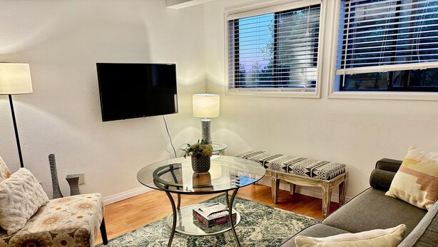 A living room with a glass coffee table, a sofa, a patterned armchair, a TV mounted on the wall, and a window with blinds.