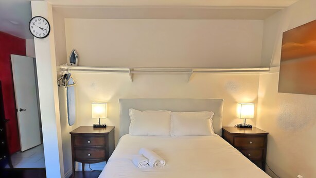 A neatly made bed with white linens and a folded towel, two bedside lamps, a clock on the wall, and a red door in the background. - MaGiQ Private Master En-Suite w Private Bathroom, No Shared Spaces (Spring Valley)