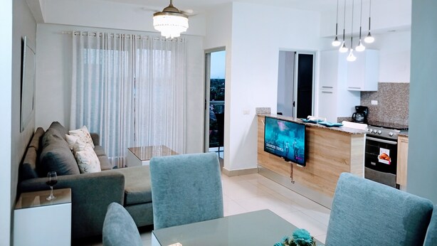 A modern living room with a sofa, chairs, a glass coffee table, and a TV displaying 'Stranger Things'. A kitchen is visible with a stove, microwave, and cabinets.