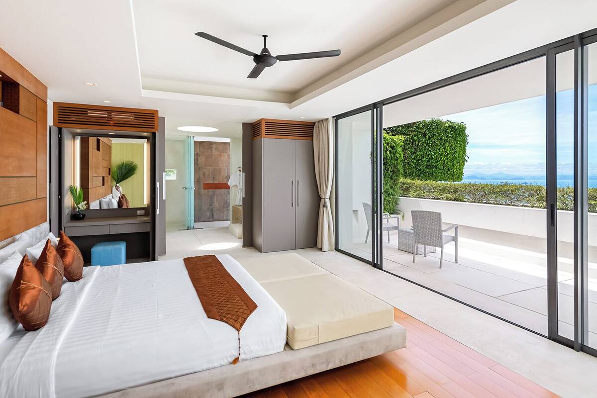 Ein modernes Schlafzimmer mit einem großen Bett, einem Deckenventilator und Blick auf einen Balkon mit Gartenmöbeln.
