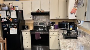 Una cocina con armarios blancos, un refrigerador negro, una estufa y un microondas. Hay una alfombra en el suelo y una silla en la esquina.