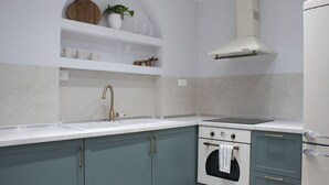 Une cuisine avec un évier blanc, une cuisinière et une hotte de ventilation blanche. On y trouve des placards sous le plan de travail et une étagère au-dessus de l’évier.