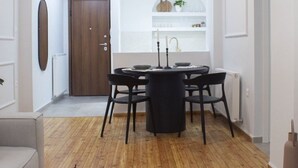 Un salon moderne avec un plancher en bois, une table de salle à manger ronde entourée de chaises noires, une table basse noire sur laquelle reposent un livre et des bâtonnets d’encens, et un mur blanc agrémenté d’une porte en bois et d’une étagère où sont disposés des objets décoratifs.