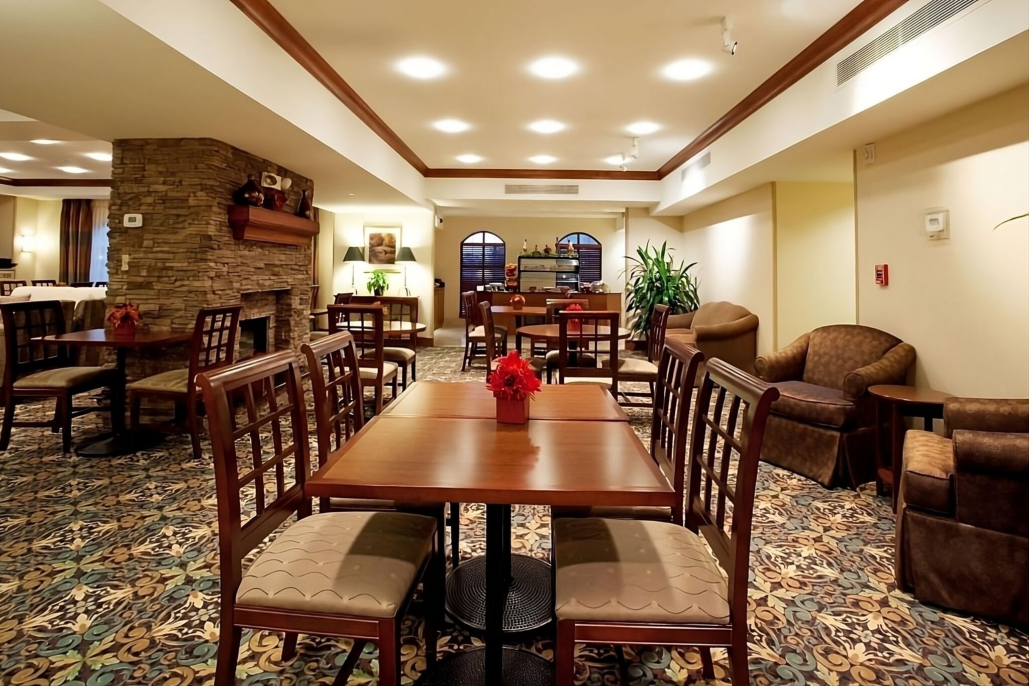 A hotel dining area with a stone accent wall, wooden tables and chairs, and a patterned carpet.