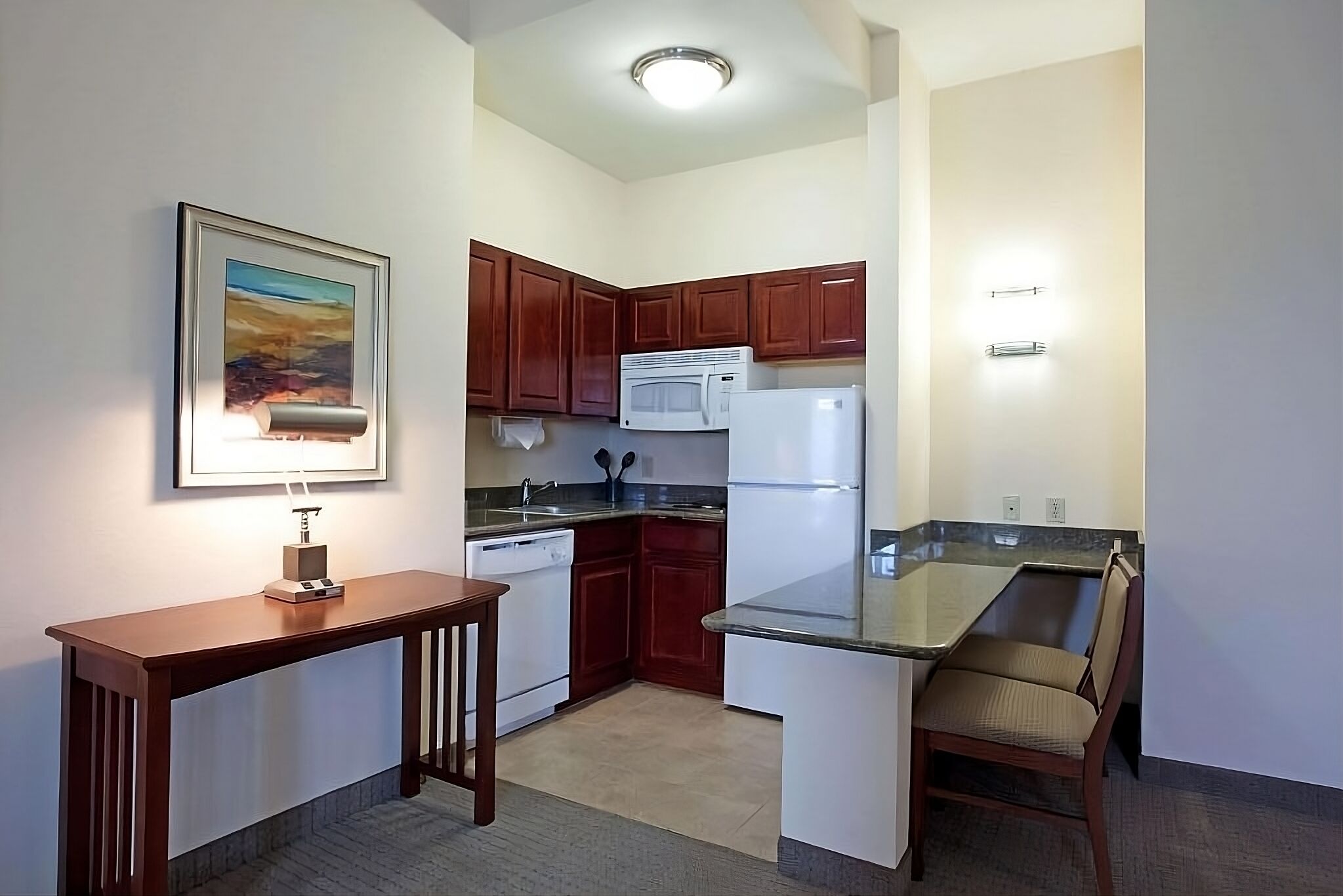 A compact kitchen with wooden cabinets, a microwave, a refrigerator, a small dining table, and a chair.