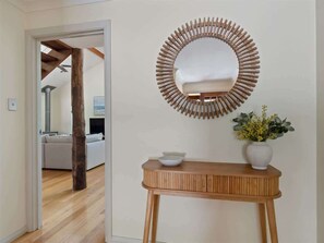 Un couloir avec une console en bois, un vase blanc orné de fleurs et un miroir rond doté d’un cadre en bois.