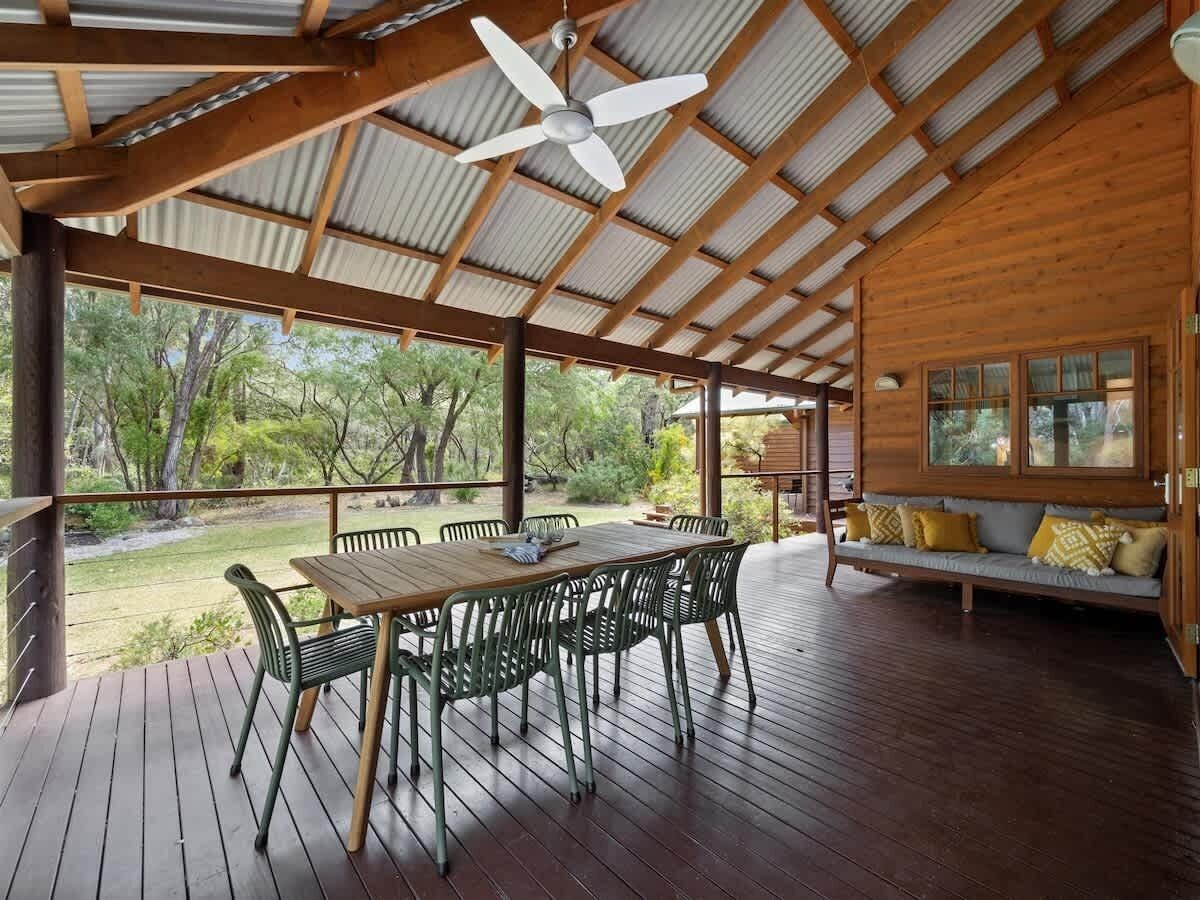 Une terrasse extérieure avec une table et des chaises en bois, un ventilateur de plafond et une vue sur les arbres environnants.
