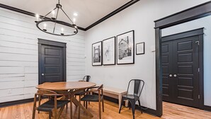 Un comedor con una mesa y sillas de madera, un banco y una araña. Hay tres cuadros en la pared y dos puertas negras.