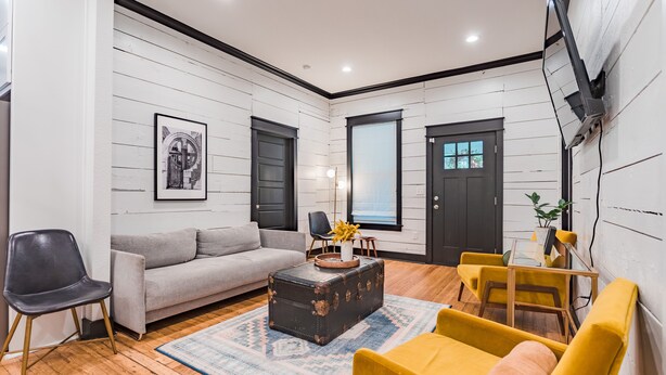 Una sala de estar moderna con un sofá gris, sillones amarillos, un baúl antiguo y un ventanal grande.