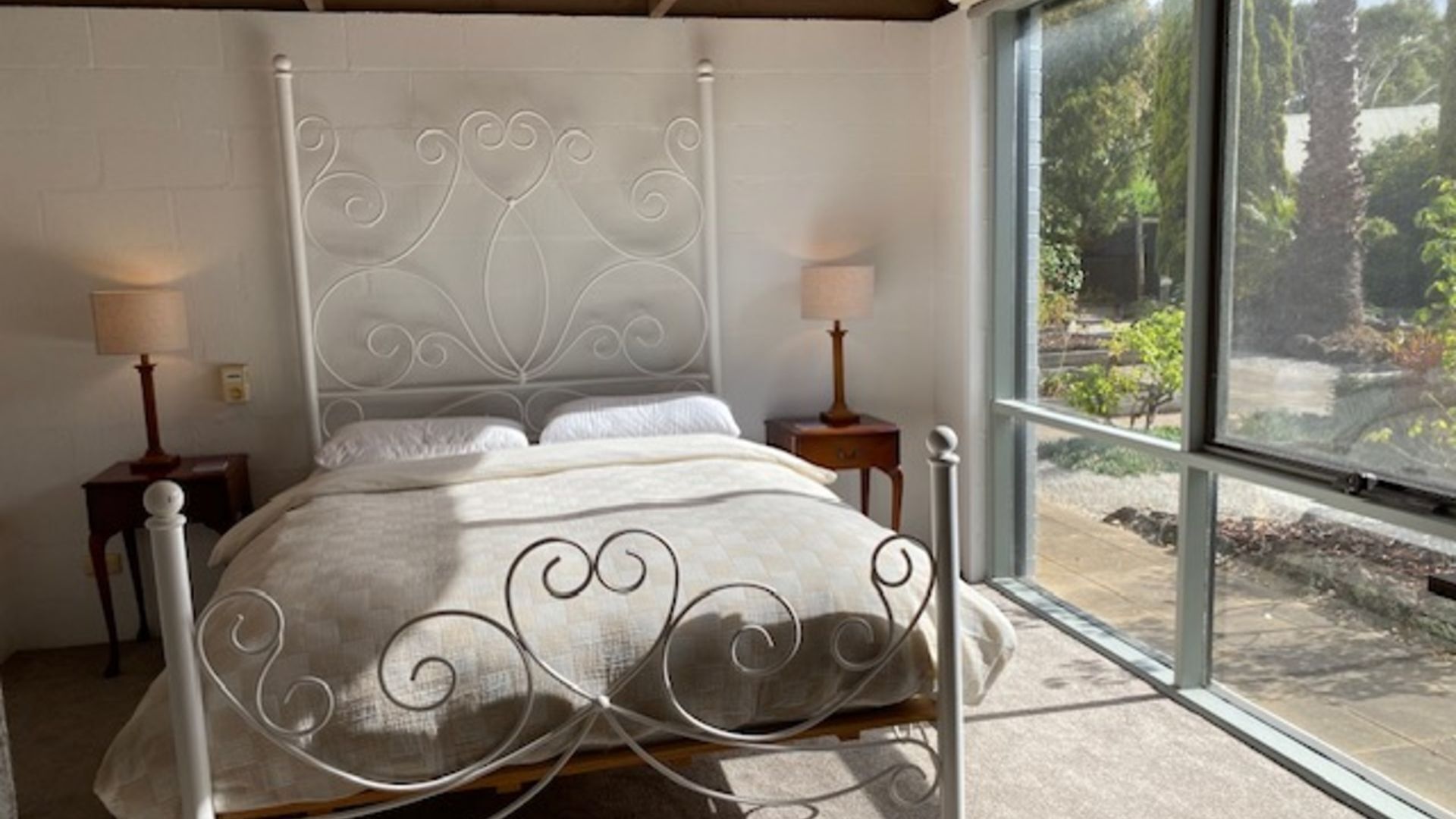 A bed with a white metal frame and a decorative headboard, two bedside tables with lamps, and a view of a garden through a large window.