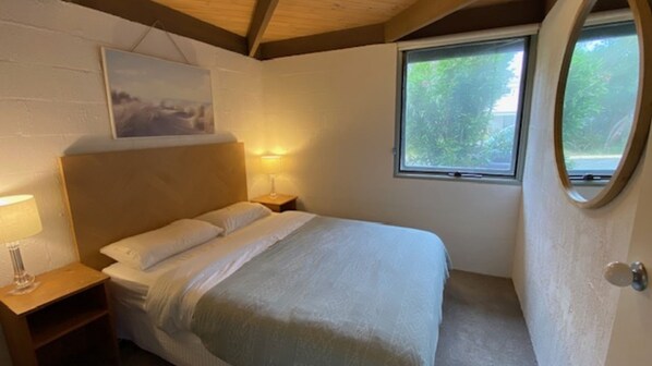 A bedroom with a bed, a nightstand, a lamp, a painting on the wall, and a window with a view of trees. - A Sand and Sea Retreat - 150m from Beach, Pet Friendly (Inverloch)