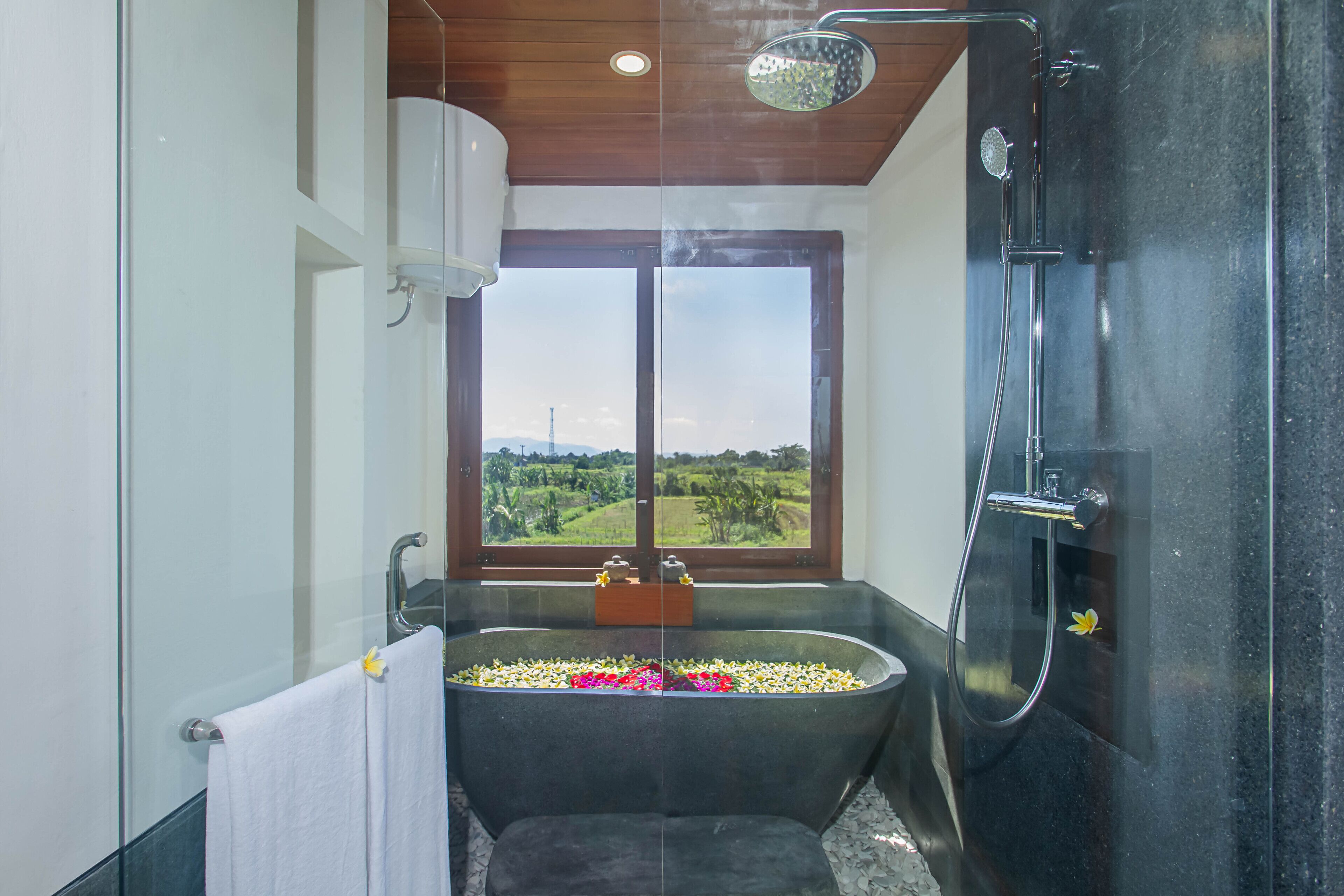 A bathroom with a bathtub filled with flowers, a shower, and a view of the countryside through a large window.